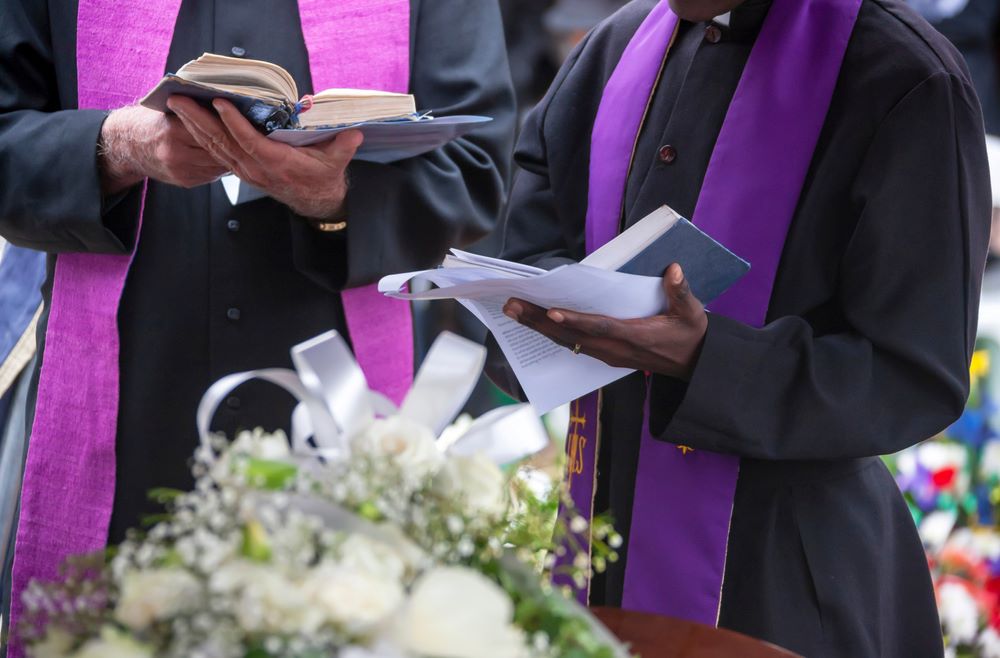 Image showing a Christian funeral service in a church, with mourners gathered around a casket adorned with flowers. The minister leads the service, offering prayers and comforting words to the grieving family and friends. The atmosphere is solemn and reflective, conveying a sense of faith, hope, and the journey towards eternal peace